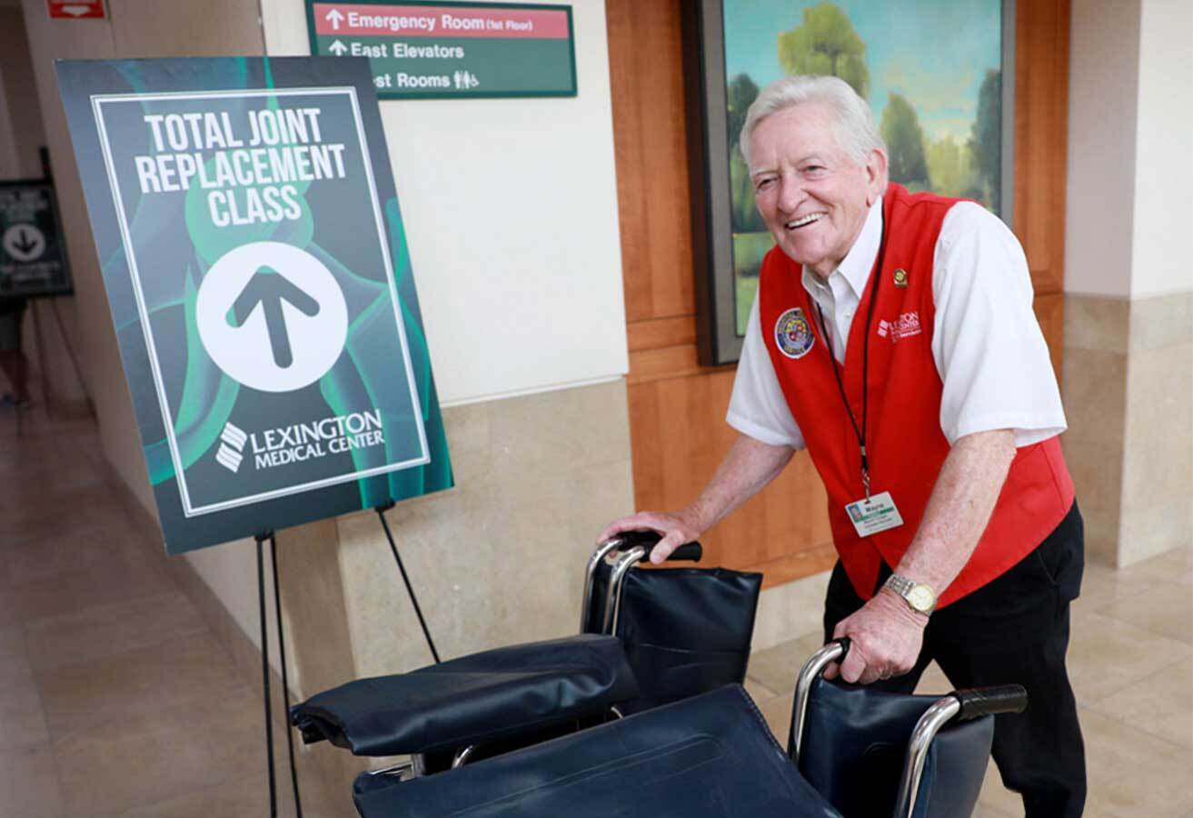 A smiling volunteer pushing two wheelchairs past a "Total Joint Replacement Class" sign.