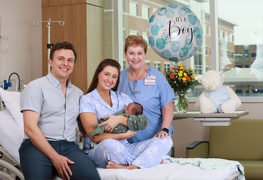 A smiling nurse and family holding a new born baby with a "It's a Boy" balloon behind them.