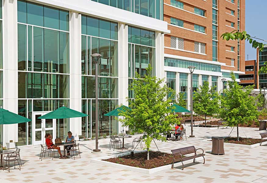 The outside courtyard at the North Tower of the hospital. There are tables and chairs covered with umbrellas and trees planted around the courtyard.
