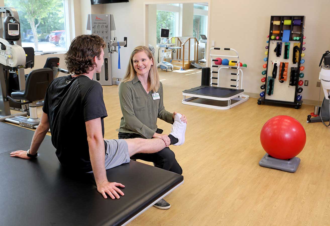 A smiling physical therapist examining a patient's foot in an office with exercise equipment.